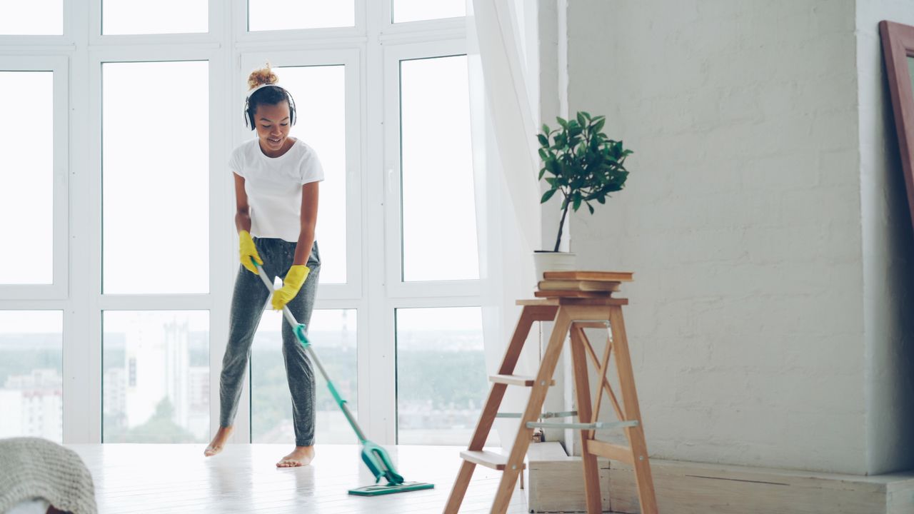 Young woman cleaning floor with a mop in a sunny room with large windows.