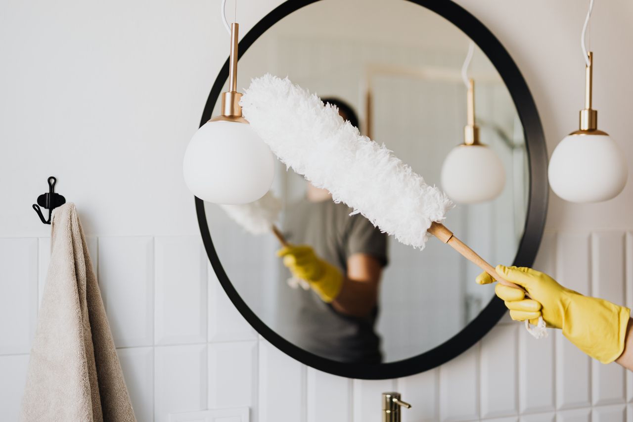 programs-02 Crop anonymous female housewife in rubber gloves dusting modern lamp with soft brush against mirror while cleaning stylish bathroom