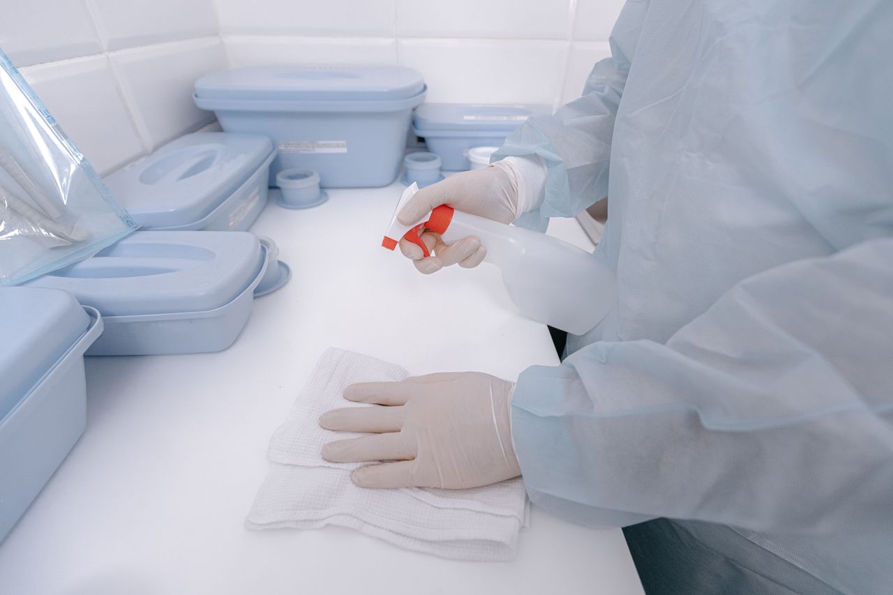 programs-03 A healthcare worker wearing gloves sanitizes a surface with a spray bottle in a medical setting.